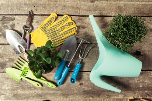 A collection of gardening tools and accessories arranged on a weathered wooden outdoor surface, including a pair of pruning shears, yellow gardening gloves, a small hand trowel, a weeding fork, a plastic watering can, and a green-handled spray nozzle, alongside a potted plant with lush green foliage. The background appears to be an outdoor garden space with visible wooden decking nearby. The tools and plant are positioned in a slightly scattered manner, suggesting preparation for gardening or outdoor maintenance in a typical garden setting that could be found in Ealing, London. The scene features natural tones of wood, green plants, and colorful garden tools, with lighting indicating daytime conditions suitable for gardening activities. The image aligns with garden maintenance, lawn care, and landscaping services provided by Landscapers Ealing, emphasizing the practical tools used in maintaining a healthy and well-kept garden or outdoor space.