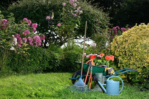 In a well-maintained garden, a patch of vibrant green grass extends across the foreground, bordered by various flowering bushes and shrubs. On the left, pink and purple rhododendrons are in full bloom, adding vivid colour to the landscape, while on the right, a large yellow-green shrub provides contrast. In the middle ground, a small flower bed is partially visible behind the shrubs, bordered with wooden edging. Several gardening tools and equipment are arranged in the garden, including a blue watering can, a rake with blue tines, and a wheelbarrow filled with gardening supplies such as gloves, cloths, and small plant pots. The garden appears to be in a residential back yard, with dense trees and greenery in the background, suggesting a private outdoor space in Ealing. The scene is lit with natural daylight under partly cloudy skies, highlighting the variety of plant textures and colours, and emphasizing the connection between garden maintenance and professional landscaping activities often provided by Landscapers Ealing.