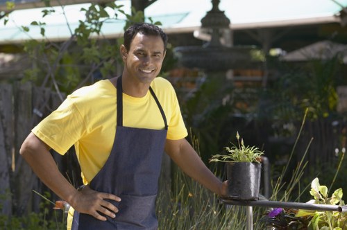 A smiling male gardener with medium skin tone, wearing a yellow T-shirt and a dark grey apron, stands outdoors in a well-maintained garden. He is holding a black plant pot containing a green, leafy plant, and is positioned near a small round table with garden tools and additional plants. The garden features a lawn with dense, healthy grass, edged by flower beds and various shrubs. In the background, there are mature trees, a wooden fence, and garden structures, suggesting a landscaped outdoor space. The scene appears to be on a bright, sunny day with natural light illuminating the area, highlighting the lush greenery and vibrant colours of the plants. This setting reflects professional gardening and landscaping services offered by Landscapers Ealing, contributing to a tidy and inviting garden environment in the Ealing area.