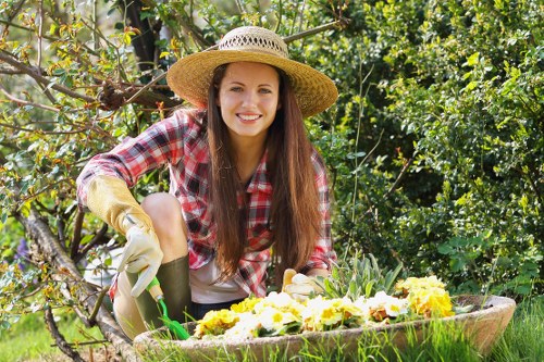 A young woman with long brown hair, wearing a wide-brimmed straw hat, a red and white checkered shirt, and gardening gloves, is kneeling in a lush garden. She is smiling while tending to a raised flower bed filled with blooming yellow and white flowers, adjacent to a well-maintained lawn with dense green shrubbery and trees in the background. The garden features a mix of grass, soil, and stone paving, with vibrant natural tones and well-organized plantings, typical of outdoor landscaping in the Ealing area. The scene is illuminated by natural daylight, indicating a clear, sunny day, highlighting the careful maintenance and growth of the garden elements. This setting supports professional gardening and landscaping practice, emphasizing outdoor horticultural care consistent with services offered by Landscapers Ealing. The garden's design includes neatly bordered flower beds, lush green hedge lines, and a combination of lawn and paved surfaces suitable for variety of outdoor gardening activities and landscaping improvements.