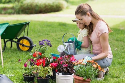 The image depicts a young woman and a child working together in a lush green garden with a well-maintained lawn, surrounded by a variety of vibrant flowering plants and potted flowers in red, purple, and orange hues. They are crouched next to a collection of gardening tools and watering can, carefully tending to the plants. In the background, there is a neatly trimmed hedge, and a green wheelbarrow with yellow handles is visible on the grass, suggesting ongoing outdoor gardening or landscaping activities. The scene is set outdoors on a bright, sunny day, with natural light illuminating the garden’s textures and colors. This scene captures a moment of outdoor gardening, emphasizing garden enhancement activities suitable for landscaping and lawn care services in Ealing, UK, and highlights the importance of plant care and garden organization, as provided by companies like Landscapers Ealing.