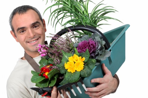 The image shows a smiling man holding a teal plastic garden trowel and a garden fork, with a round black metal tray filled with colorful flowering plants including purple and yellow blooms, inside a garden space with lush greenery in the background. The garden features a well-maintained lawn with dense, dark green grass, bordered by a neat hedge and a paved patio area. The environment appears bright and outdoor, suggesting a sunny day, and the scene reflects professional gardening work, possibly provided by local landscapers such as Landscapers Ealing. The overall setting highlights a tidy, landscaped garden with a variety of flowers and lush foliage, typical of a residential outdoor space in the UK, maintained to a high standard suitable for gardening and landscaping services.