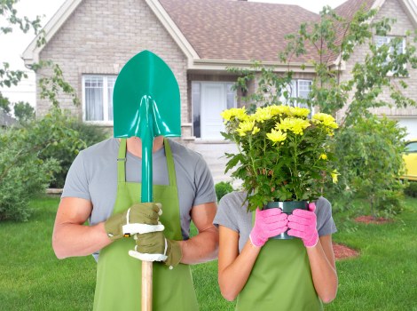 A gardener and a woman, both wearing green gardening aprons and brightly coloured gloves, are standing in a well-maintained front garden with a lush green lawn, surrounding shrubs, and a small flower bed. The gardener holds a green-handled shovel with a shiny metal blade, while the woman is holding a large pink pot filled with vibrant yellow flowering plants. In the background, there is a modern brick house with white window frames and a grey tiled roof, framed by trees and bushes. The scene suggests outdoor gardening work in a residential setting during daylight hours, with natural light highlighting the fresh, natural colours of the plants and surroundings, reflecting professional gardening and landscaping services typical of Ealing or nearby West London communities.